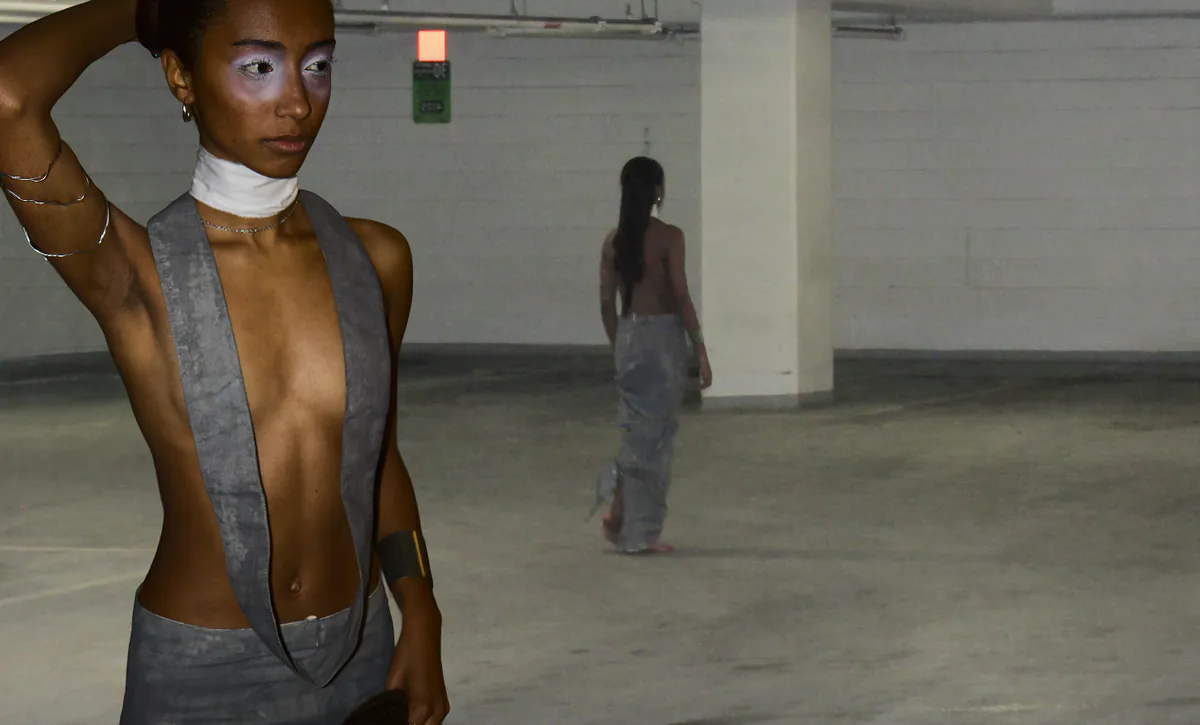 A model in a grey draped vest and white neck wrap stands in a parking garage while a second model walks away behind her.