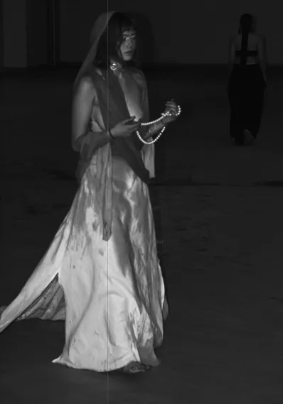 A model in a long pale dress holding a beaded strand, lit in the dark garage.