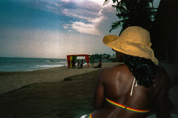 A woman in a straw hat looking out to sea, a red, gold and green canopy on the sand.