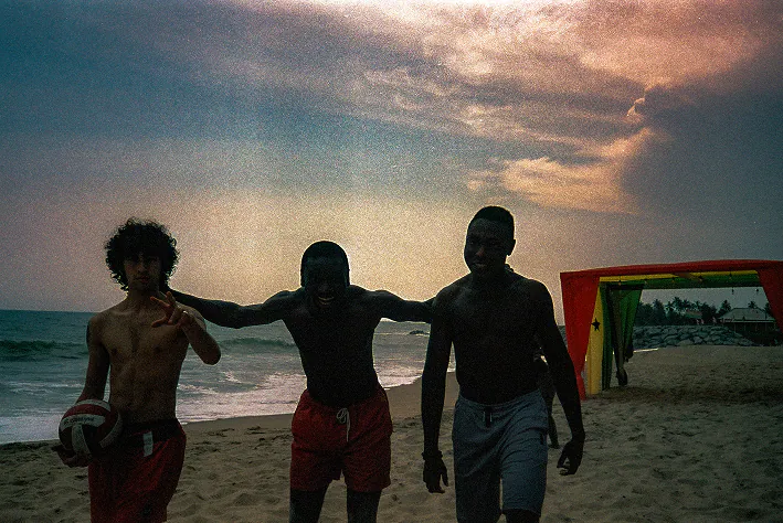Three young men with their arms around each other on the beach at dusk.