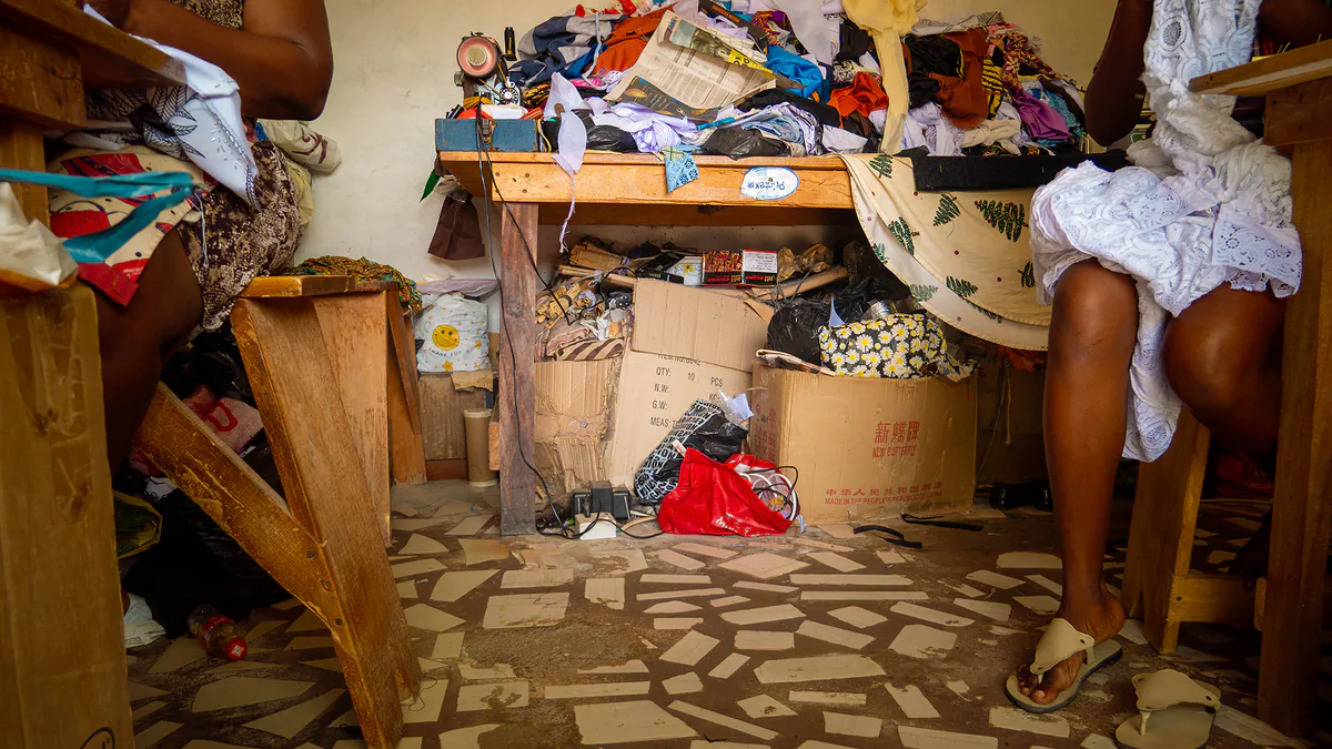 Two people seated either side of a stall piled with clothes and fabric.