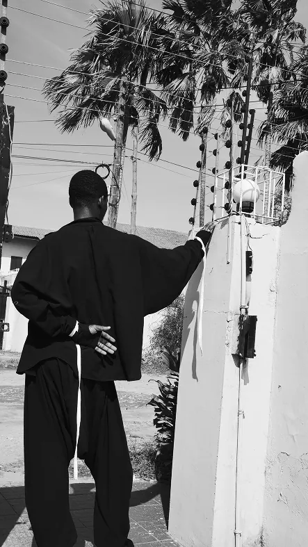 Black and white: a man seen from behind in a loose black jacket and ballooning trousers, one hand on a gatepost, palm trees and power lines overhead.