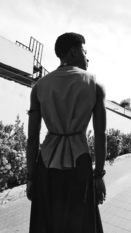 Black and white: a man from behind in a pale sleeveless vest tied at the waist, the bare back of his shoulders in sunlight.