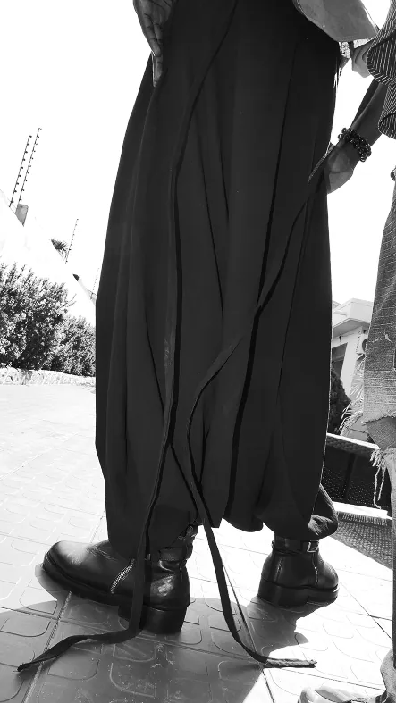 Black and white, from the ground: long black ties trailing from the trouser hem down to heavy black boots.