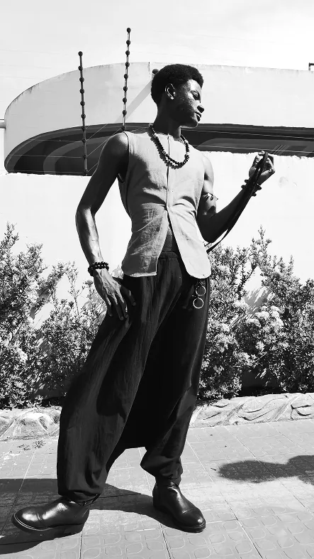 Black and white: a man in the pale vest, a beaded necklace and full black trousers, posing on a paved walk under a curved white canopy.