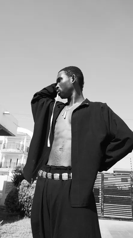 Black and white: a man in an open black jacket and beaded belt, one hand on his head, against a bright sky.