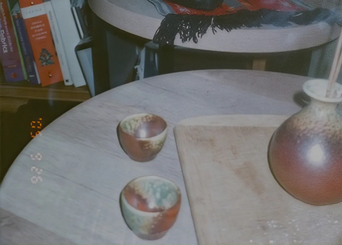 Two small ceramic cups and a vase on a round wooden table, a film date stamp in the corner.