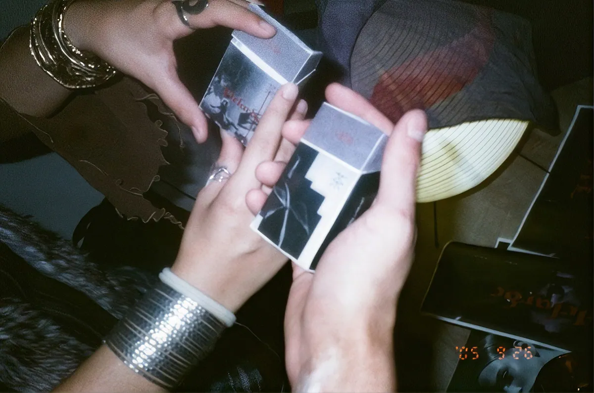 Two hands holding cigarette-box style tea packs in a dim room.