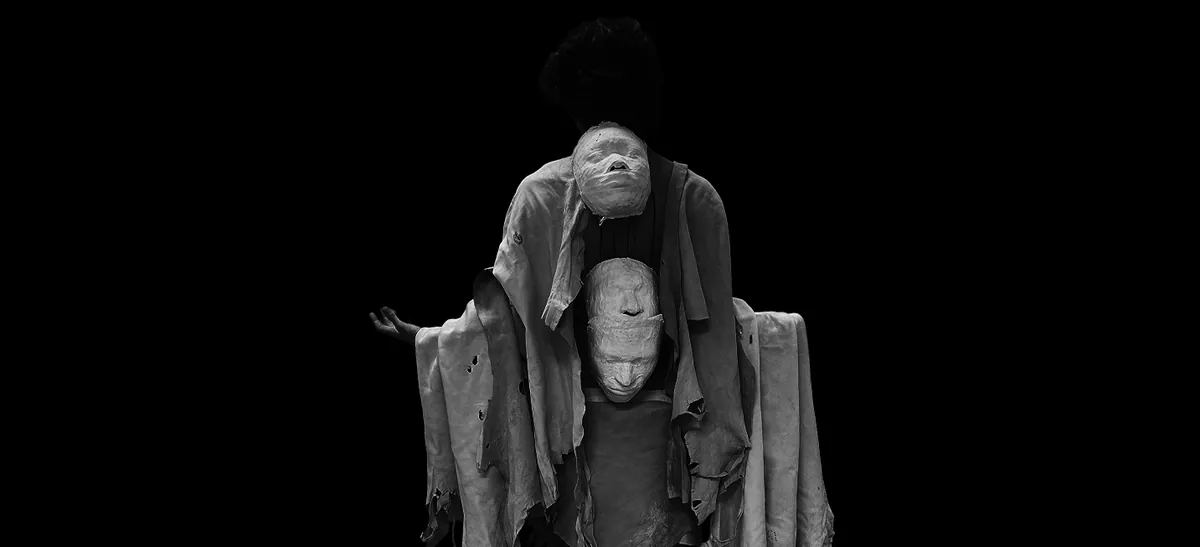 Two stacked plaster faces on a figure draped in rough cloth, against black.