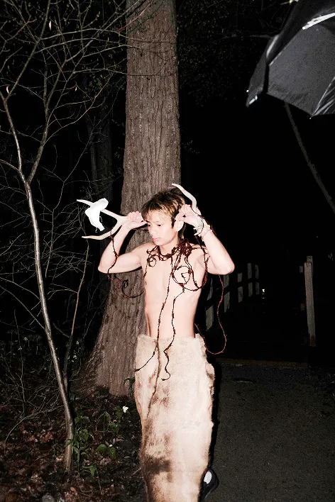 A performer in antlers and loose cords standing by a tree at night.