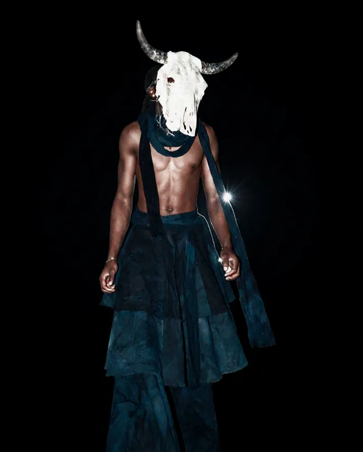 A model in a skull mask and layered indigo garments, lit by flash.