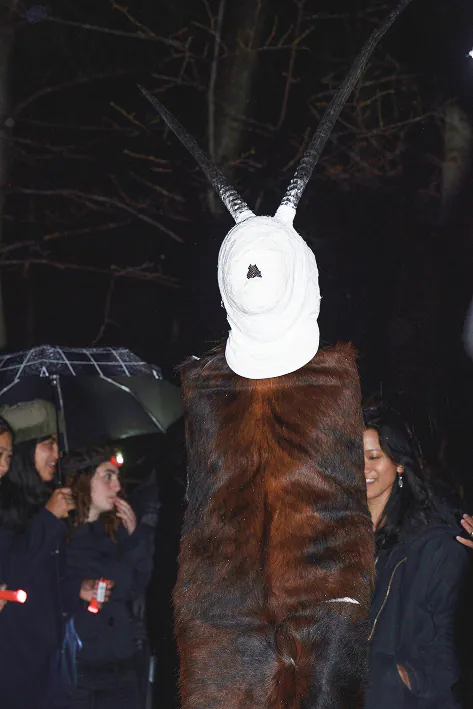 A white horned mask and fur cape among guests holding umbrellas.